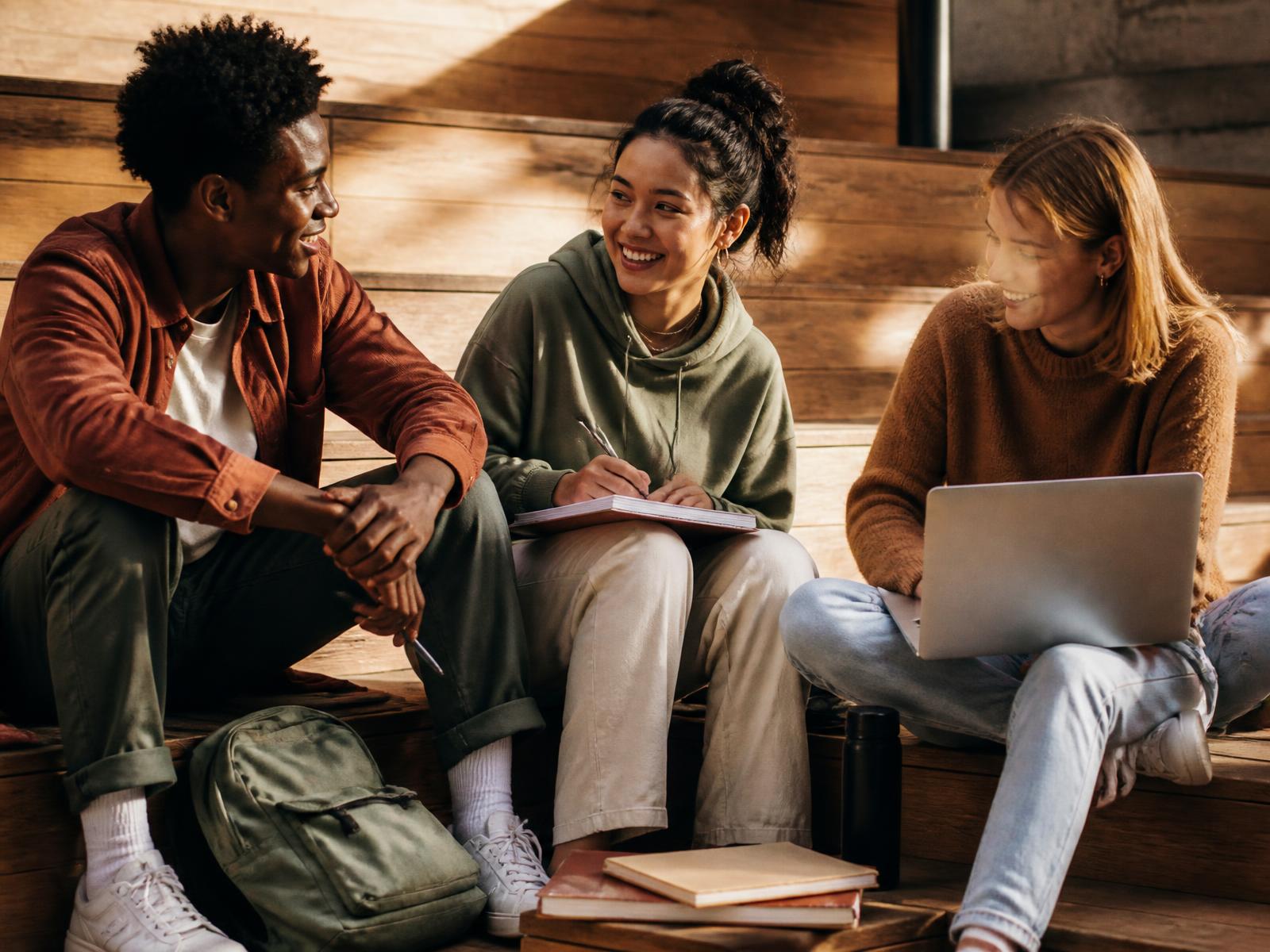 Three students sitting on wooden steps talking, with notebooks and a laptop